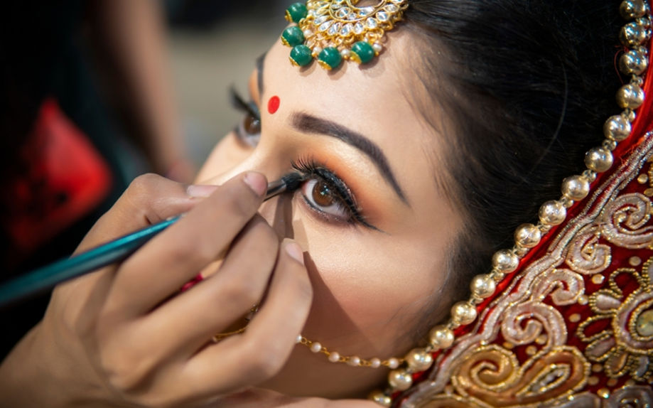 A close-up of a bride's eye makeup being done, with traditional jewelry, a red bindi, and colorful attire.