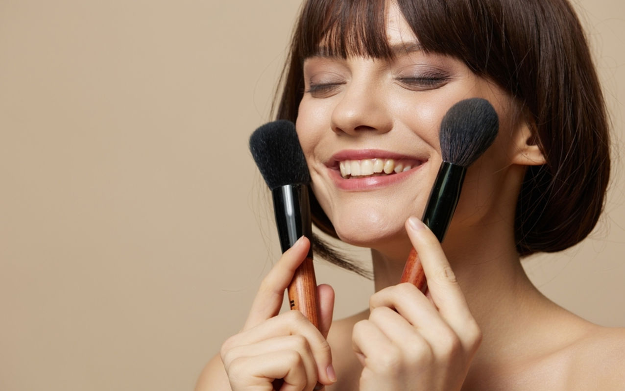 A smiling woman holds two makeup brushes to her cheeks, eyes closed, enjoying a beauty moment.