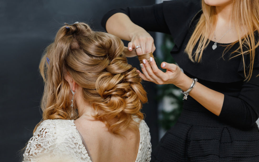 A hairstylist is creating an elegant updo on a blonde woman, using pins and styling tools, with detailed curls.